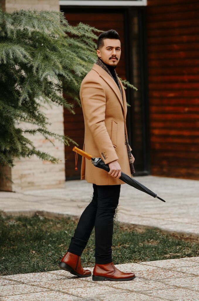 A man in a smart camel coat, black pants and brown shoes walks along a footpath with an umbrella