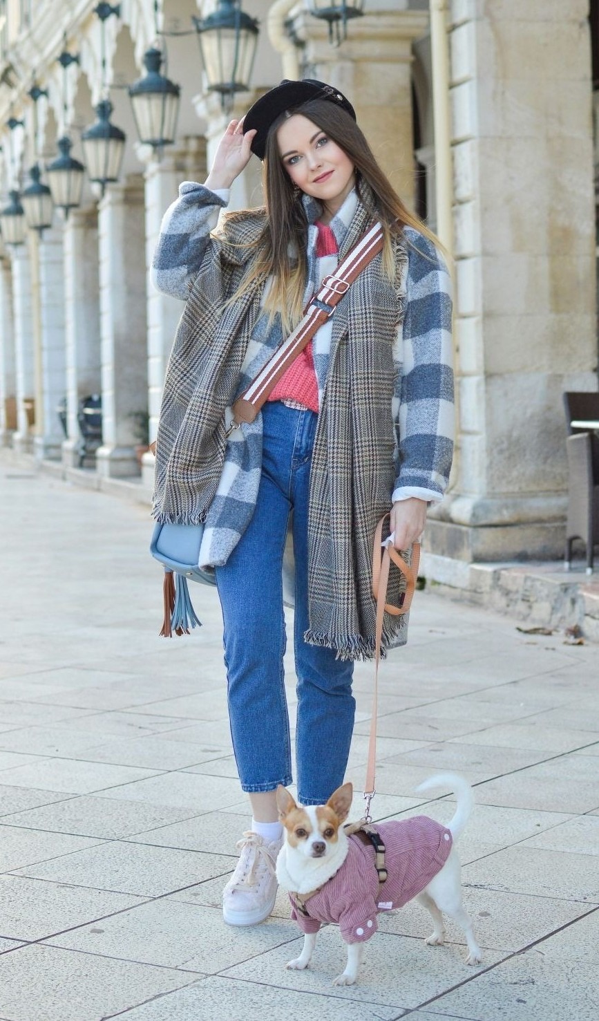 A young woman dressed casual-chic in cropped blue jeans, a coral sweater, blue striped overcoat, long plaid scar, black beret and pink sneakers poses while walking her chihuahua at Liston Square in Corfu (Greece)