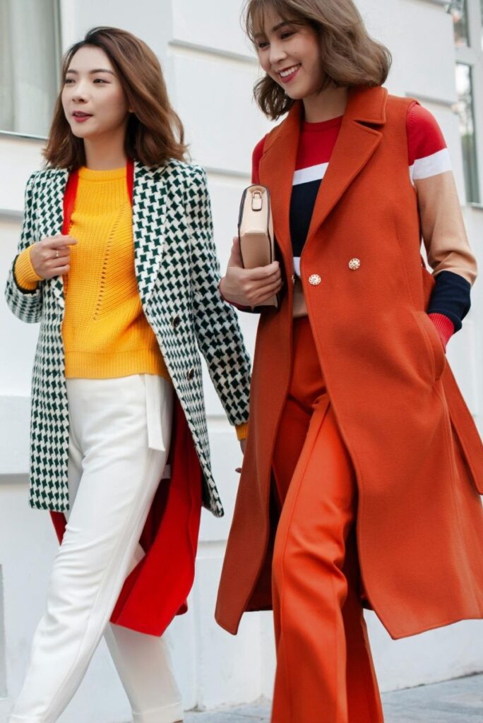 Two women walk down a footpath stylishly dressed. On the left, woman wears a sunflower yellow sweater with white pants under a black and white chevron-style jacket and long tomato red cardigan. On the right, woman wears a chic burnt orange sleeveless knee-length suit jacket and matching pants over a rust red, white, tan, and black block-colour striped sweater