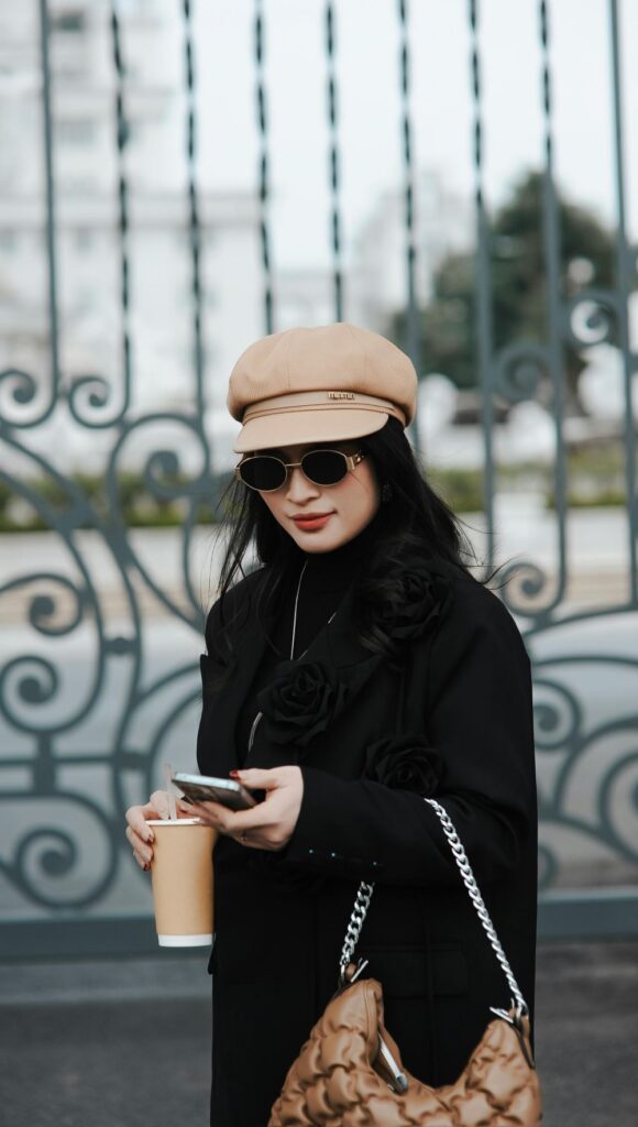 A woman texts on her phone with one hand while holding a takeaway drink in the other in front of a wrote iron gate wearing a chic camel hat, sunglasses, and a black overcoat with a tan bag