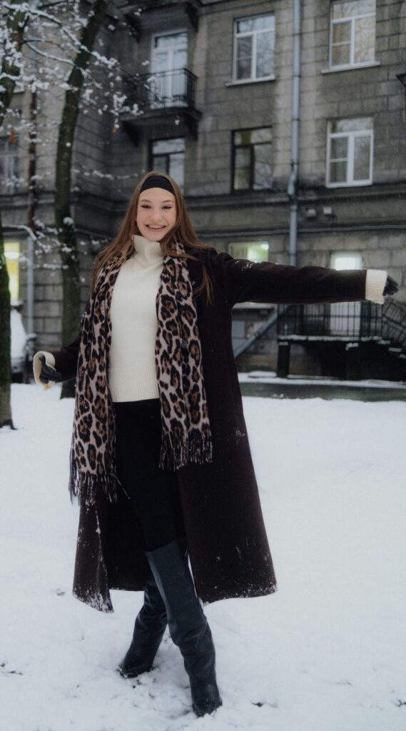 A woman smiles and poses in the snow in front of an apartment building wearing a warm cream turtleneck, long black overcoat, black headband, and hanging leopard-print scarf - warm, playful and stylish February fashion