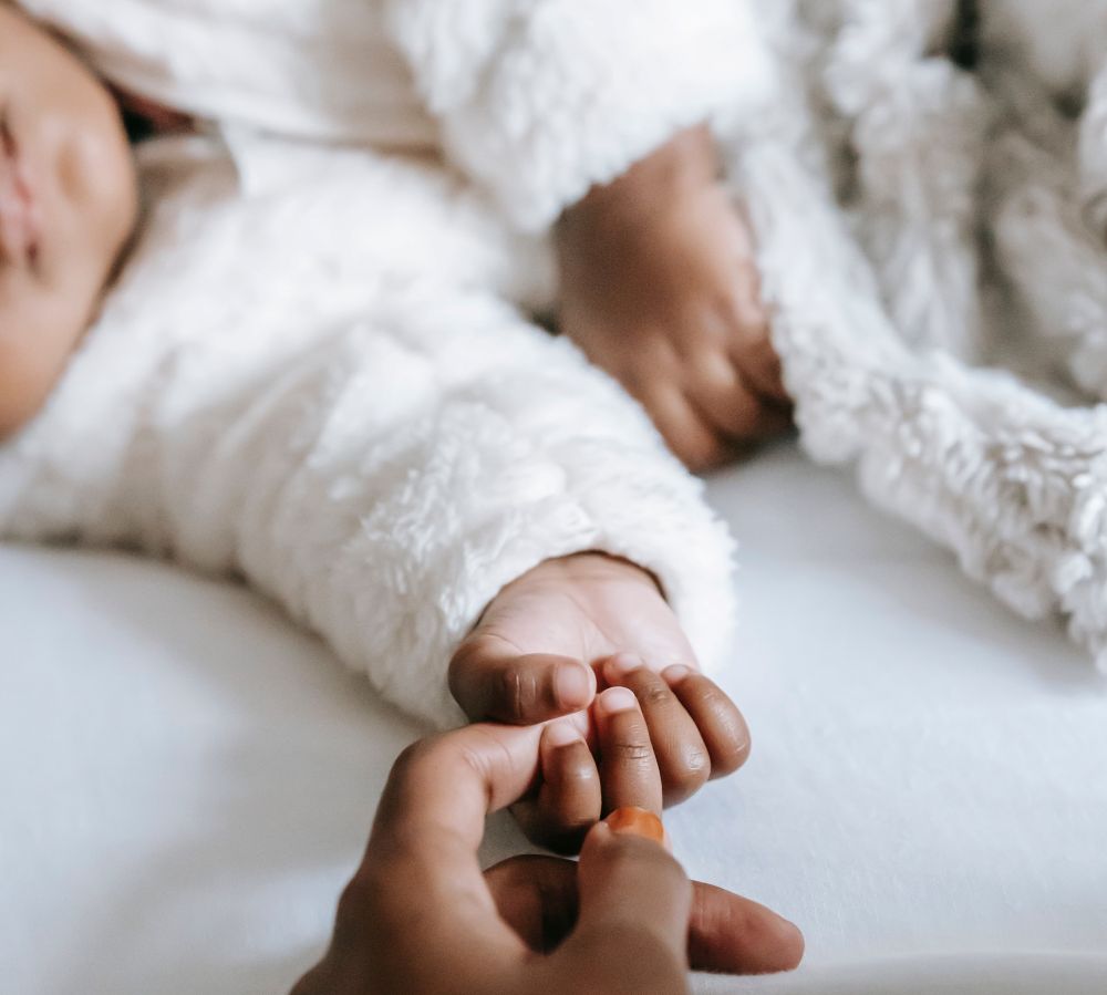 A sleeping baby with her arm held out clasps mom's finger in her hand