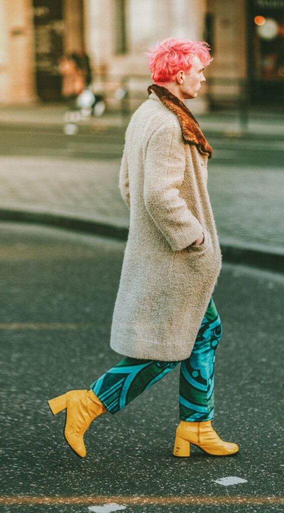 A man with cotton candy-pink hair crosses a London street in a long beige coat with dark orange-lined lapel, blue and green patterned pants, and bright yellow patent leather heeled boots