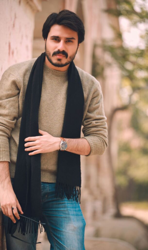 A man wears blue jeans, a greige crewneck sweater and a black scarf