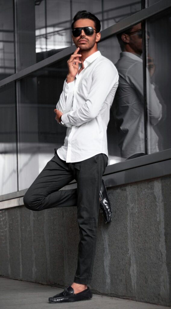 A man stands against a wall in Tehran wearing a white button-up collared shirt, black sunglasses, classy black pants and black shoes