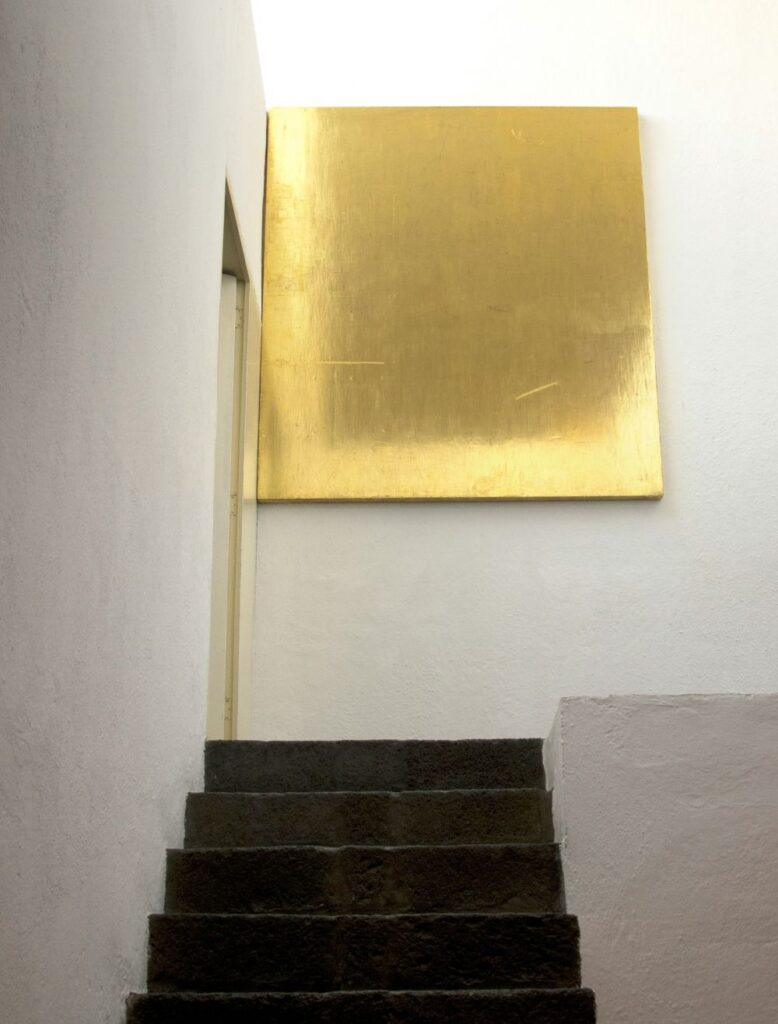 Photo showing concrete stair steps between white walls leading up to a landing with a large bright shining minimalist gold decor wall art hanging in Ciudad de México