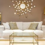 A room with gold decor including a cream sofa behind gold-framed glass coffee table, gold sunburst mirror on wall above it, gold throw pillows, gold floor lamp and gold-framed glass end table