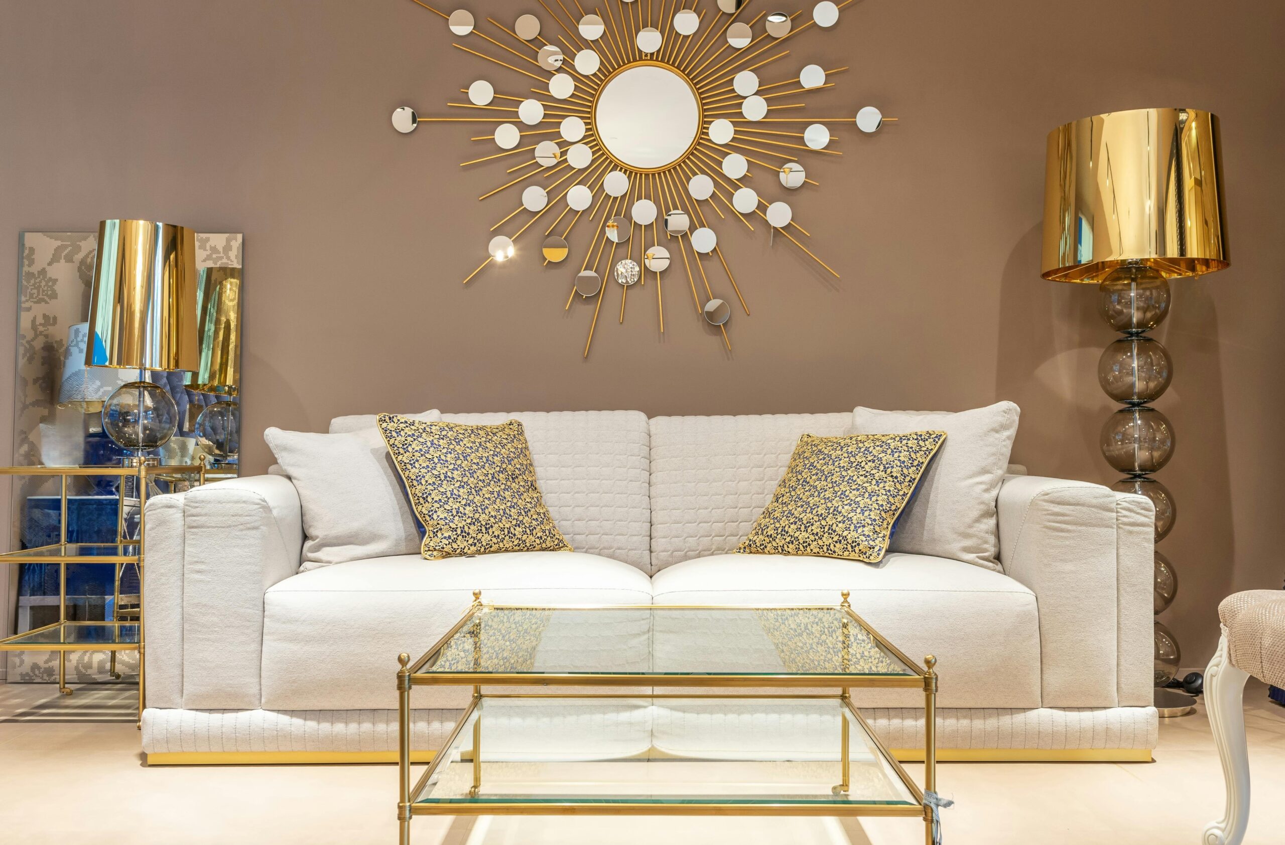 A room with gold decor including a cream sofa behind gold-framed glass coffee table, gold sunburst mirror on wall above it, gold throw pillows, gold floor lamp and gold-framed glass end table