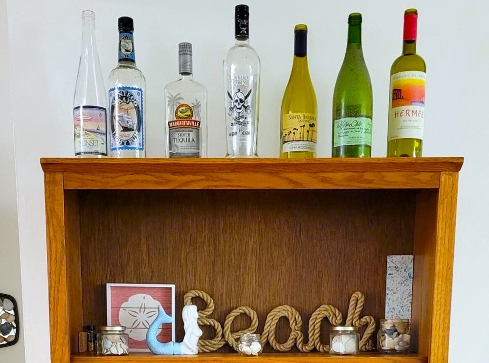 Close-up photo of the top shelf of a brown bookcase displaying coastal-themed decorations (a natural woven 'Beach' decoration, a ceramic mermaid, a small sand dollar print, and small jars of seashells). On top of the bookcase are nautical-themed wine and liquor bottles. Fun bottle labels make decorating on a budget easy