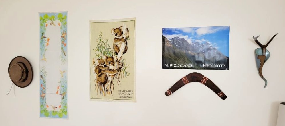 Photo of a wall with various decor: a brimmed hat from Australia with an emu feather, a silk scarf with a leaf and hummingbird pattern, a woven cloth tea towel with koalas, a poster of mountains in New Zealand, an Aboriginee boomerang,