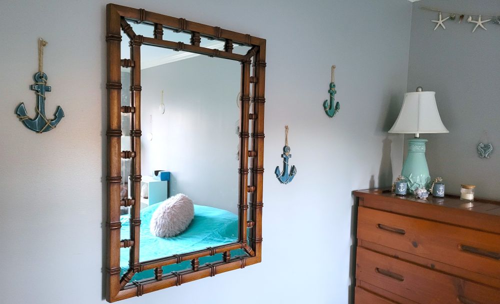 A corner of a bedroom is shown with a brown dresser, table lamp with a light blue ceramic base with sculpted sea stars, a few small bottles with anchors on them, a seahorse decoration hanging on the wall, a garland with sea stars and shells hanging above near the ceiling, and a few small blue-painted wooden anchors hanging on the wall near a large mirror on the wall with a wooden frame