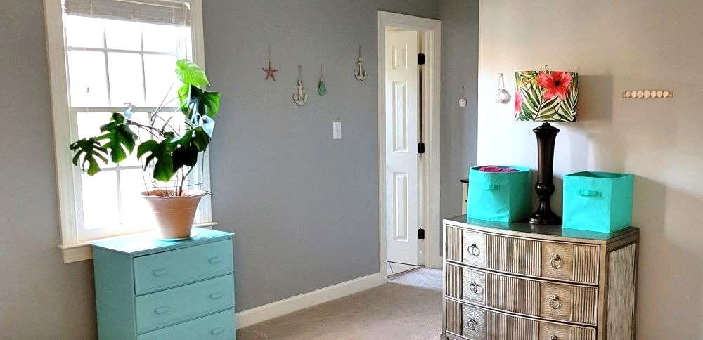 Bedroom with bright aqua-painted dresser and cloth bins, lamp with tropical fern and flower-patterned shade, monstera plant, and small seashell/aquatic decorations on wall