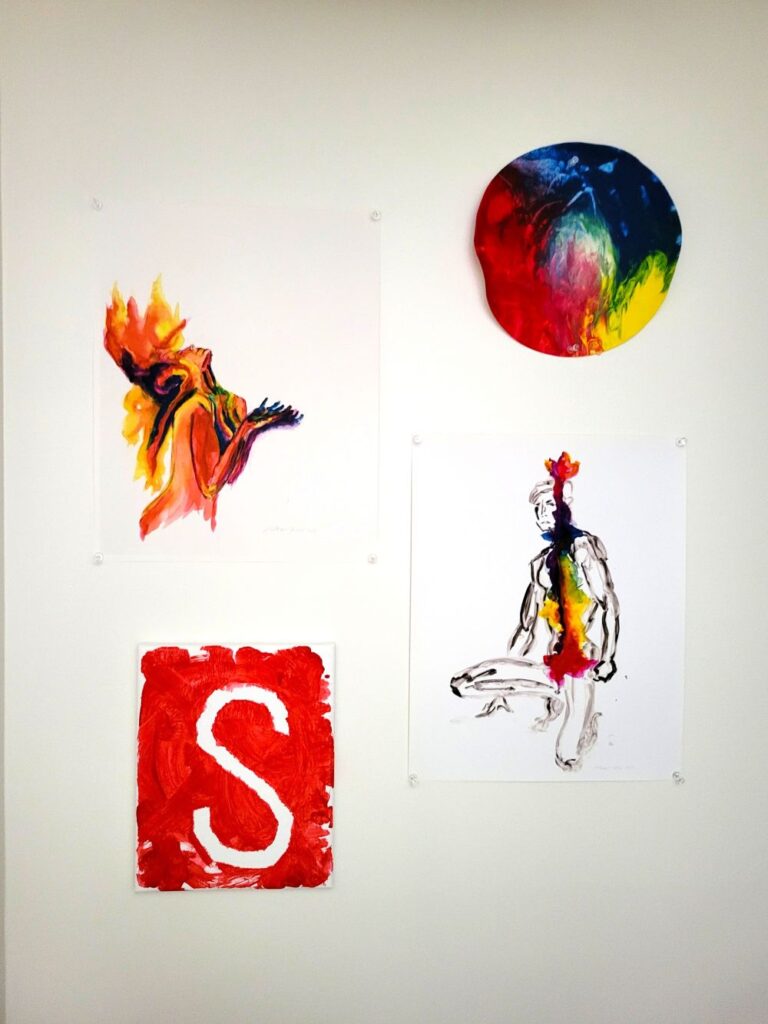 Four colourful red-centric paintings on a white wall: a round red, yellow and blue painting showing the intersection of the three primary colours, a rectangular canvas in the opposite corner showing red paint with an outline of the white letter 'S', and two professional prints of paintings of figures in blank ink coloured in with a rainbow palate