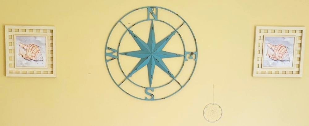 Two framed seashell prints flank a large blue metal compass rose, with a small white/clear dreamcatcher below it to the right