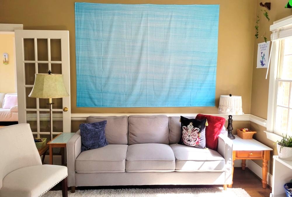 A minimalist light blue ombre lightweight tapestry hangs on a living room wall above a gray couch, offering a colour pop. When decorating on a budget, these lightweight tapestries are easy and effective ways to transform a space at a fraction of the cost of a thicker woven cloth tapestry
