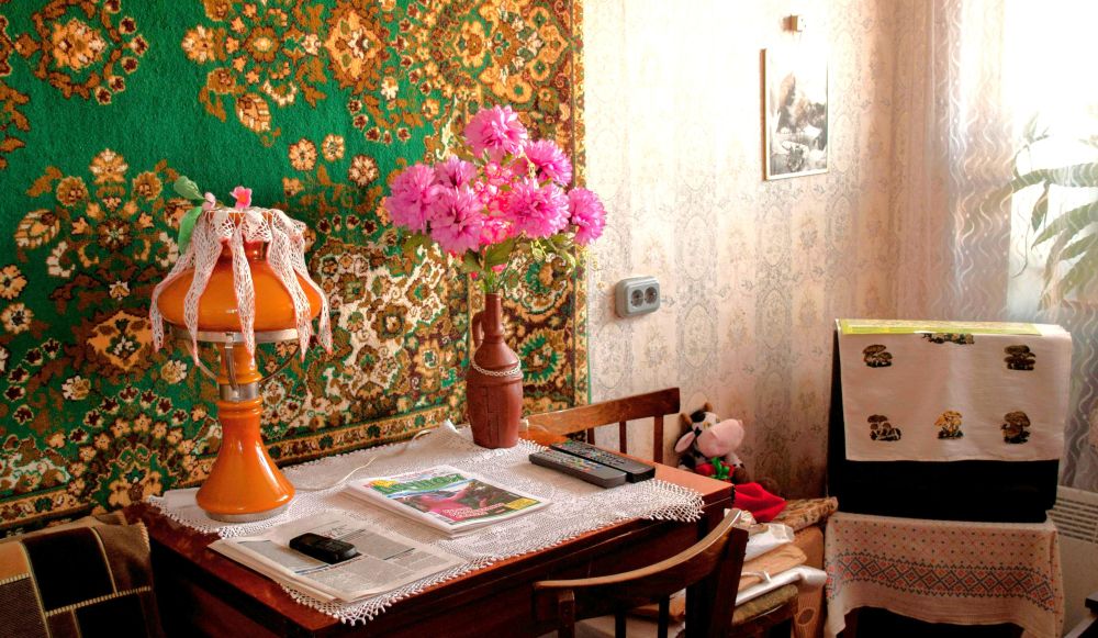 A room decorated with flower-centric prints: a flower-print traditional woven tapestry rug hangs on the wall behind a small wooden table with a white eyelet cloth atop it as well as a small table lamp adorned with matching eyelet cloth strung down the sides. A small wooden vase with a bouquet of pink flowers also sits atop the table. Nearby are a couple of chairs with hand-embroidered cloths hanging from them, and behind a sheer white curtain is a large potted plant/tree in front of a window with light streaming in. On the faint/light-coloured flower-patterned wallpapered wall hangs an indistinguishable framed piece of art