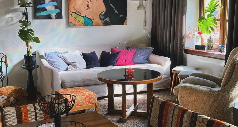 A living room with a coffee table and a few small decorative items, a few chairs, stools, monstera plants, modern art paintings/prints on the wall, and an off-white sofa covered in six throw pillows of various colours