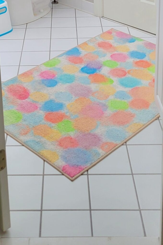 A colourful bathroom rug with multiple light/pastel coloured splotches covers part of a white-tiled bathroom floor