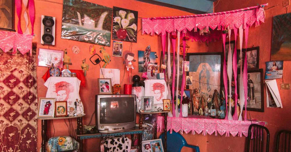 Colourful maximalist room with religious decor in Coyolillo, Mexico: in front of bright coral-coloured walls are a corner shrine outfitted in embellished pink fabric and hanging streamers with many religious photos and icons, many photographs of children and people, pictures of flowers, small images, a television on a small table, hanging patterned fabric, and chairs