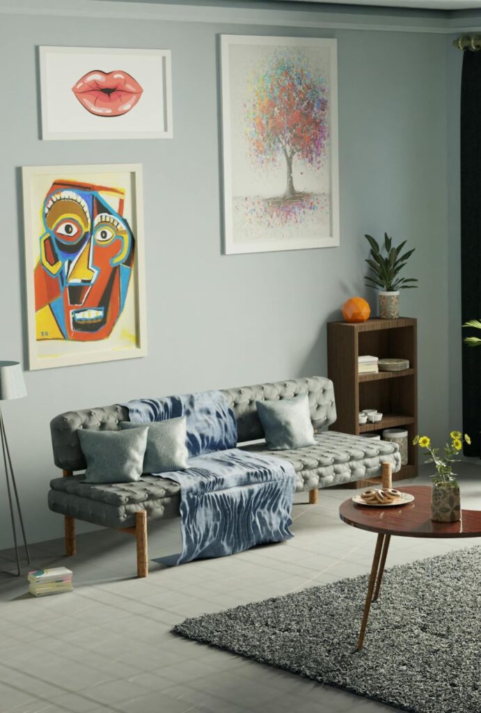Living room with modern wall art in Dhaka, Bangladesh. Three large framed prints of colourful and modern designs (abstract face, lips, and colourful tree) hang on the wall above a minimalist gray tufted couch with shiny gray-blue throw pillows and a blue throw blanket. On a small shelf beside the sofa is a small potted plant and round orange object. On a coffee table over a textured gray rug is a plate of cookies and a small vase with yellow flowers