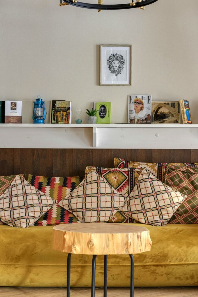 A mustard sofa is strewn with various patterned throw pillows. In front of it is a small round wooden table, and above it is a shelf on the wall displaying a small plant, an hourglass, a lantern, and various books and magazine. A minimalist drawing of a lion hangs on the wall above it