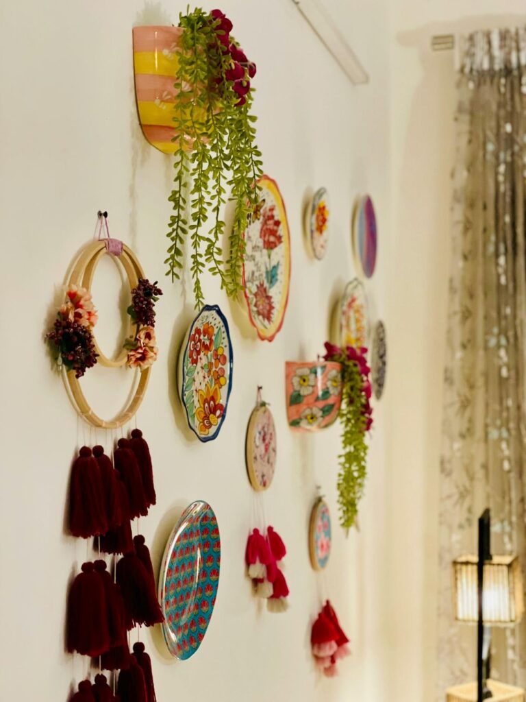 Close-up photo of dainty and colourful decorations hanging on a wall: a hoop with small flower adornments and multiple hanging maroon tassels, a small pink and yellow-striped painted vase with a small sedum plant with red flowers hanging out of it and another nearby, other tasseled decorations, and several colourful painted decorative plates