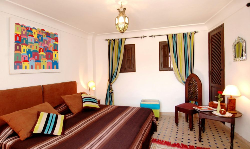 A Moroccan-style bedroom furnished with a brown striped blanket over a king bed with brown and yellow, white, blue, and brown striped throw pillows, matching curtains and footstool, a colourful painting/print hung on the wall above, and other small decorative touches