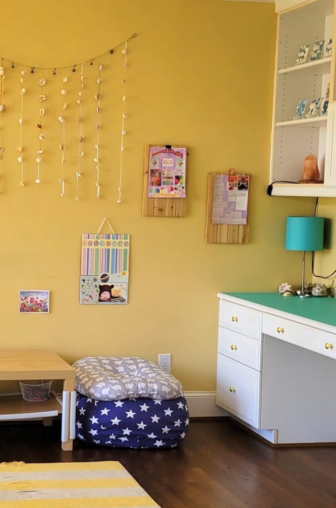 Decorating on a budget with a creative flair: a room with a yellow wall, yellow and white-striped rug covering part of the floor, built-in white desk with teal peel-and-stick wallpaper covering the top surface, a matching aqua lamp under shelves with hand-made decorations and a salt lamp, hand-crafted seashells on ribbons hanging on the wall, a few hand-made collages hanging on the wall, and a white-spangled blue plush/pillow/bedding case stuffed with blankets and a pillow on top next to a coffee table