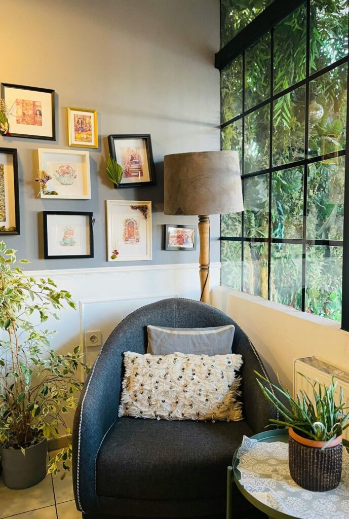 A small upholstered chair sits in the corner of a room next to a large window with foliage outside and a floor lamp behind it. On the wall behind it is a gallery of framed pictures, and to the other side is a small end table with a potted plant. On the floor nearby is a larger potted plant. On the chair are a couple of textured throw pillows. Plants and throw pillows are perfect for decorating on a budget