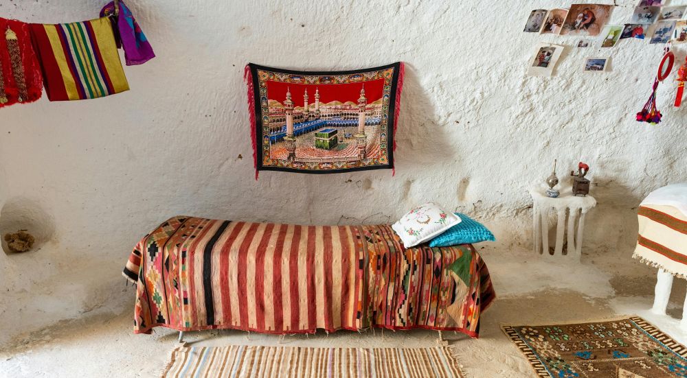 An underground house in Matmata, Tunisia is decorated with a traditional print blanket over the bed, a traditional woven rug on the floor to the right, another striped rug on the floor in front of it, a small table to the right with a woven rug over it, small decorations on a built-in small table (a natural outcropping carved out of the wall), many photographs hung on the wall above it along with colourful pompom decorations, and a clothesline with colourful fabrics hanging from it on the other side. Above the bed is an exquisitely-detailed woven tapestry/rug of the Ka'bah in Mecca in bold colours