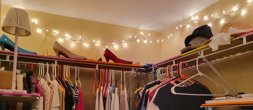 White string lights are strung across the ceiling inside a closet, illuminated a row of different-coloured pumps as well as some hats atop the shelf above hanging clothes
