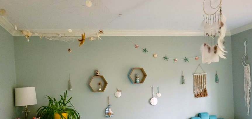 View of the upper half of a bedroom with a decorative fishing net hanging in the corner with starfish and seashells hanging in it, a white-feathered dreamcatcher hanging from another part of the ceiling, and many nautical-themed decorations hanging on the wall.