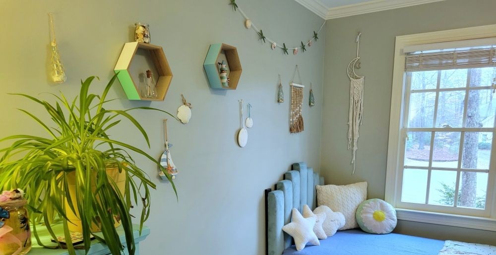 Nautical-themed decor in light blue hues hang from a bedroom wall with a sailboat, seashells, sand dollars, and more. A cream-coloured macrame crescent moon-shaped dreamcatcher hangs on the adjacent wall, too